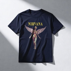 May include: Navy blue t-shirt featuring the word "NIRVANA" in yellow block letters above a graphic of a nude figure with feathered wings. The figure has a detailed anatomical design.