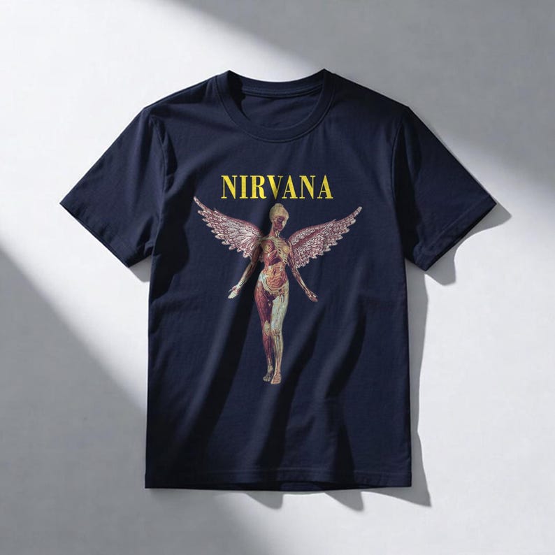May include: Navy blue t-shirt featuring the word "NIRVANA" in yellow block letters above a graphic of a nude figure with feathered wings. The figure has a detailed anatomical design.