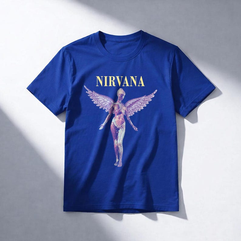 May include: Royal blue t-shirt featuring the word "NIRVANA" in gold lettering above a graphic of a nude figure with angel wings. The figure has a detailed anatomical design. The shirt is a classic crew neck style.