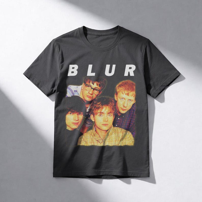 Puede incluir: Camiseta gris oscuro con la palabra "BLUR" en letras blancas sobre una imagen de estilo vintage de cuatro hombres. La camiseta est&aacute; hecha de un material suave y es adecuada para uso casual.