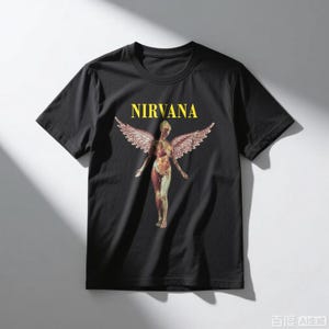 May include: Black t-shirt featuring the word "NIRVANA" in yellow block letters above a graphic of a nude figure with angel wings. The figure is in a standing pose with arms outstretched. The shirt is made of a soft material.