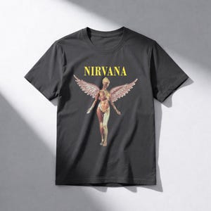 May include: Dark gray t-shirt featuring the word "NIRVANA" in yellow block letters above a graphic of a nude figure with angel wings. The figure's internal anatomy is visible. The shirt is a classic crew neck style.