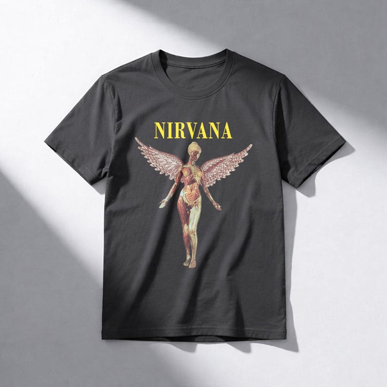 May include: Dark gray t-shirt featuring the word "NIRVANA" in yellow block letters above a graphic of a nude figure with angel wings. The figure's internal anatomy is visible. The shirt is a classic crew neck style.