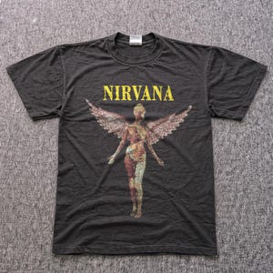 Peut inclure: T-shirt gris foncé avec le mot "NIRVANA" en jaune au-dessus d'un graphique d'une figure nue avec des ailes d'ange. L'anatomie interne de la figure est visible. Le t-shirt est à manches courtes.