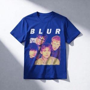 Puede incluir: Camiseta azul real con la palabra "BLUR" en letras blancas. Debajo, una imagen descolorida de la banda, con cuatro hombres. La camiseta es de estilo cuello redondo cl&aacute;sico.