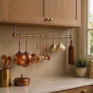 Puede incluir: Una barra de cocina de latón montada bajo armarios de madera, con ganchos en S que sostienen sartenes de cobre, un tenedor, una cuchara, un cucharón, dos tazas de cerámica color crema y una tabla de cortar de madera.