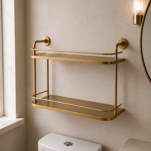 May include: A two-tier wall-mounted shelf with a brushed gold finish. The unit features two rectangular shelves with rounded corners, each enclosed by a thin metal railing. It is secured to the wall with two circular mounting brackets at the top.
