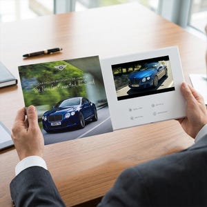 May include: A person holding a white video brochure featuring a blue Bentley Continental GT Speed car. The brochure displays the text "YOUR EXCLUSIVE INVITATION TO THE LAUNCH OF The New Continental GT Speed". The video screen shows a blue car.