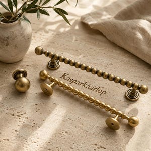 May include: Two brass-toned metal drawer pulls with a beaded texture and a single matching round knob. The pulls feature a long, slender bar design with spherical accents along the length. The text "Kasparkaarlop" is visible between the two pulls.
