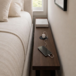 May include: A dark brown wooden bedside table with a built-in charging station and a smartphone plugged in. A book is on the table. The table is positioned between a bed and a wall.