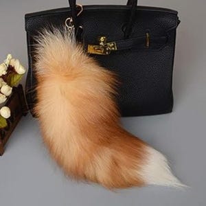 Peut inclure: Un porte-clés en forme de queue de renard orange et blanc duveteux suspendu à un sac à main en cuir noir. La queue présente un dégradé de couleurs, passant d'un orange clair à une pointe blanche. Le sac à main a des détails dorés.