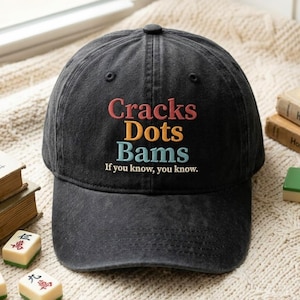 May include: A black baseball cap with embroidered text in red, yellow, and blue. The text reads Cracks, Dots, Bams, and below it, If you know, you know. The cap features a curved brim and a structured crown with ventilation eyelets on the top panels.