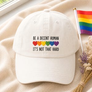 May include: A white baseball cap featuring black embroidered text that reads BE A DECENT HUMAN IT'S NOT THAT HARD, positioned above a row of six small embroidered hearts in red, orange, yellow, green, blue, and purple colours.