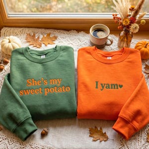 Könnte beinhalten: Ein waldgrüner Pullover mit dem orangefarbenen Sticktext 'She's my sweet potato' und ein leuchtend orangefarbener Pullover mit dem grünen Sticktext 'I yam' und einem kleinen grünen Herzsymbol, beide mit Rundhalsausschnitt.