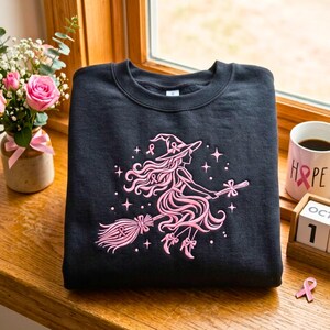 以下が含まれることがあります： ほうきに乗って飛ぶ魔女のピンクの刺繍が施された黒いクルーネックのスウェットシャツ。デザインには、なびく髪、とんがり帽子、小さな星のアクセントが含まれています。魔女の帽子とほうきには、小さなピンクのリボンが飾られています。