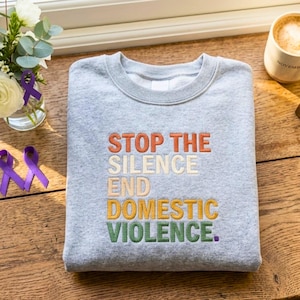 Peut inclure: Un sweat-shirt gris clair à col rond avec un texte brodé dans un design empilé et multicolore. Les mots STOP THE SILENCE END DOMESTIC VIOLENCE sont inscrits en fils orange, crème, beige clair, jaune et vert avec un point final violet.