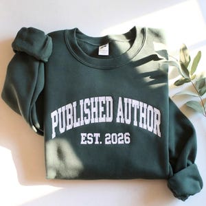 Puede incluir: Una sudadera de cuello redondo en color verde bosque oscuro con texto bordado en blanco que dice PUBLISHED AUTHOR en una fuente universitaria arqueada, con el texto EST. 2026 centrado justo debajo en una tipografía de bloque blanca.
