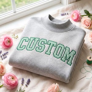 Puede incluir: Una sudadera gris claro con cuello alto y la palabra CUSTOM bordada en el pecho con un patrón de cuadros vichy verde y blanco con un borde verde oscuro. Las letras están en una fuente de estilo universitario, negrita y en bloque.