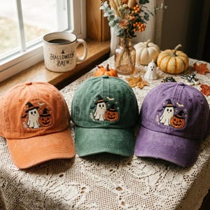 May include: Three baseball caps in orange, green, and purple, each featuring an embroidered design of a ghost wearing a witch hat next to a jack-o'-lantern. The caps have a distressed fabric texture and a curved brim, showcasing a seasonal theme.
