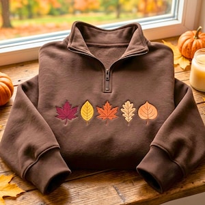 May include: A dark brown quarter-zip sweatshirt featuring an embroidered row of five distinct autumn leaves in shades of deep red, yellow, orange, beige, and light brown across the chest. The garment has long sleeves with ribbed cuffs.