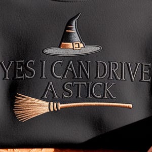 May include: A black sweatshirt featuring an embroidered design of a black witch hat with a gold band and a tan broomstick. The text "YES I CAN DRIVE A STICK" is embroidered in black lettering between the hat and the broomstick on the front.