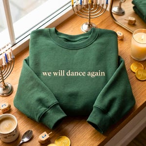 May include: A forest green crewneck sweatshirt featuring the text "we will dance again" printed in a light cream colour across the chest. The garment has long sleeves with ribbed cuffs and a ribbed neckline, presented in a flat, folded position.
