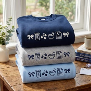 May include: A stack of three crewneck sweatshirts in navy blue, heather gray, and light blue. Each sweatshirt features a white or dark blue graphic print of a bow, a prescription bottle, two pills, a mortar and pestle, and a prescription pad.