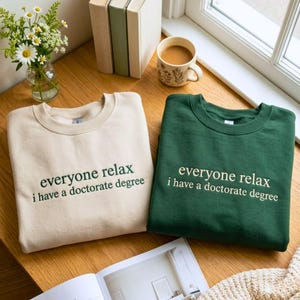 May include: A beige sweatshirt with dark green text reading "everyone relax i have a doctorate degree" and a dark green sweatshirt with beige text reading "everyone relax i have a doctorate degree" displayed on a wooden surface.