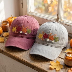 May include: A maroon and a gray baseball cap, each featuring an embroidered design of colorful autumn leaves, small berries, and white daisy flowers on the front panel. Both caps have a structured crown, a curved brim, and stitched eyelets.