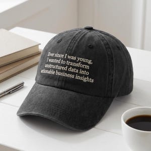 Op de afbeelding: Donkergrijze baseballcap met de witte tekst: "Ever since I was young, I wanted to transform unstructured data into actionable business insights". De cap ligt op een wit oppervlak.