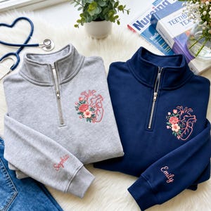 May include: A light gray and a navy blue quarter-zip sweatshirt, each featuring an embroidered anatomical heart with pink flowers on the chest and a name embroidered on the sleeve cuff, specifically Sophia on the gray and Emily on the navy.