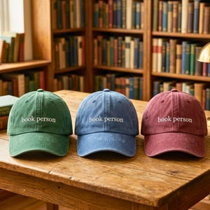 May include: Three baseball caps in green, blue, and maroon, each featuring the text book person embroidered in white on the front. The caps have a structured crown, a curved brim, and a washed fabric texture, displayed side by side on a wooden surface.
