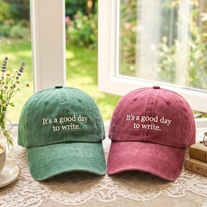 May include: Two baseball caps, one in a muted green and one in a deep rose colour, both featuring a six-panel construction with a curved brim and white embroidered text on the front that reads, It's a good day to write.