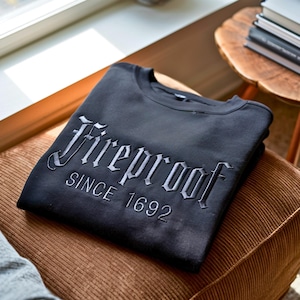 Könnte beinhalten: Ein schwarzes Sweatshirt mit Rundhalsausschnitt, das auf der Brust das Wort Fireproof in einer gotischen Schriftart gestickt hat, wobei der Text SINCE 1692 direkt darunter in einer klaren, serifenlosen Schrift für einen markanten Look sorgt.