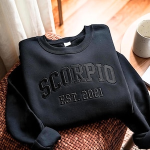 以下が含まれることがあります： 胸元に大きくアーチ状の浮き彫り文字で「SCORPIO」と書かれ、そのすぐ下に小さく同じ浮き彫りフォントで「EST. 2021」と記された黒い長袖のスウェットシャツ。