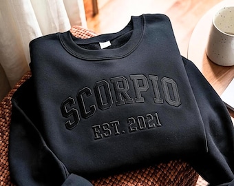 May include: A black long-sleeve sweatshirt featuring the word SCORPIO in a large, arched, raised font across the chest, with the text EST. 2021 positioned directly underneath in a smaller, matching raised font style.