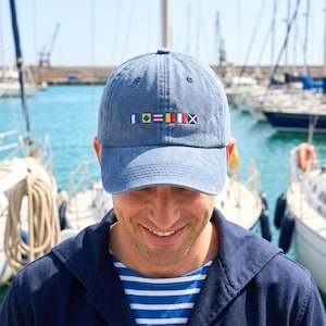 May include: A blue denim baseball cap featuring a row of six embroidered nautical signal flags across the front panel. The cap has a curved brim and a structured crown, displaying a series of colorful geometric flag patterns in a horizontal line.