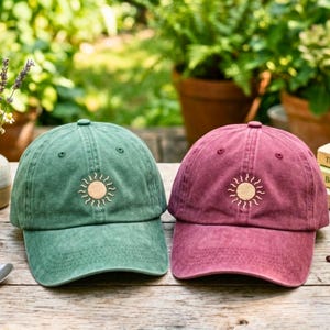 Op de afbeelding: Twee baseballcaps, één in gedempt groen en één in donkerrood, elk voorzien van een centraal geborduurd zonpictogram met een cirkelvormig midden en stralen. De caps hebben een gestructureerde kroon, een gebogen klep en ventilatiegaatjes.
