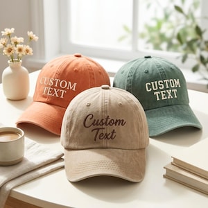 May include: Three baseball caps in orange, beige, and green. The orange and green caps feature the text CUSTOM TEXT in a serif font. The beige cap features the text Custom Text in a script font. Each cap has a curved brim and stitched panels.