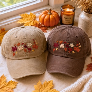 May include: Two baseball caps, one in tan and one in dark brown, each featuring embroidered autumn leaves in shades of red, orange, and cream. The caps have a curved brim and a six-panel construction with stitched eyelets on the crown.