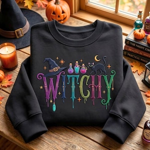 May include: A black crewneck sweatshirt featuring a colorful graphic with the word WITCHY in dripping letters. The design includes a witch hat, three potion bottles, a black cat, a crescent moon, and stars in shades of purple, blue, and green.