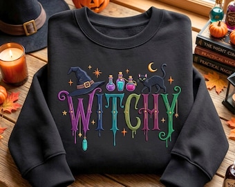 May include: A black crewneck sweatshirt featuring a colourful graphic with the word WITCHY in dripping letters. The design includes a witch hat, three potion bottles, a black cat, a crescent moon, and stars in shades of purple, blue, and green.