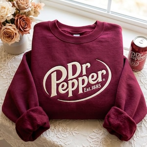 May include: A maroon crewneck sweatshirt featuring the white embroidered Dr Pepper logo and the text Est. 1885 on the front. The long-sleeved garment has a ribbed collar, cuffs, and hem, showcasing a classic pullover design with a soft texture.