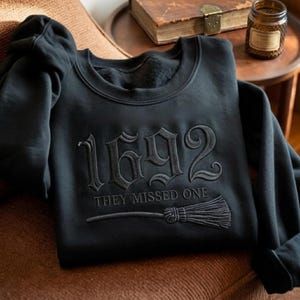 Könnte beinhalten: Ein schwarzes Langarm-Sweatshirt mit tonaler schwarzer Stickerei. Das Design umfasst den Text 1692, den Schriftzug THEY MISSED ONE und eine Grafik eines Besens. Die Stickerei ist mittig auf der Brust des Kleidungsstücks platziert.