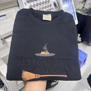 May include: A black crewneck sweatshirt featuring an embroidered design of a witch hat and a broomstick. The text "YES I CAN DRIVE A STICK" is embroidered in black thread across the centre. The hat is black with a tan band, and the broom is tan.