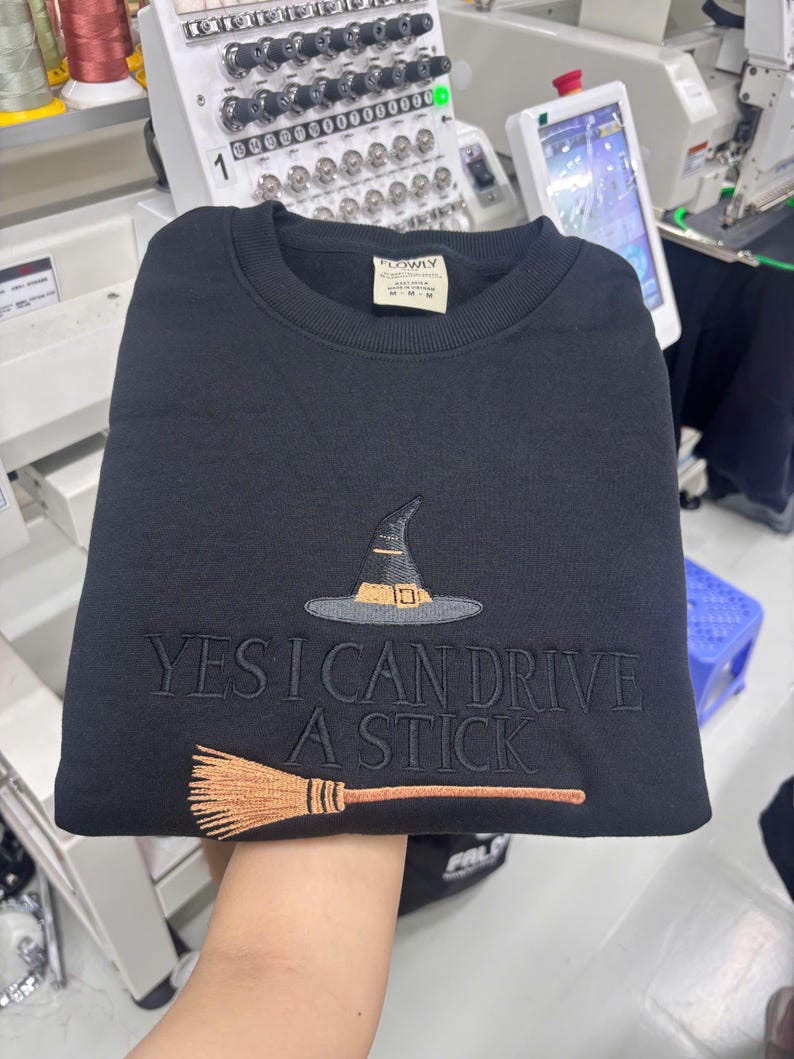 May include: A black crewneck sweatshirt featuring an embroidered design of a witch hat and a broomstick. The text "YES I CAN DRIVE A STICK" is embroidered in black thread across the centre. The hat is black with a tan band, and the broom is tan.