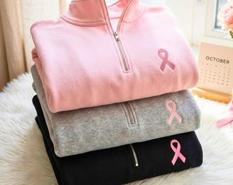 May include: A stack of three quarter-zip sweatshirts in light pink, heather grey, and black. Each sweatshirt features a silver-toned metal zip at the collar and an embroidered pink ribbon symbol on the upper left chest area of the garment.