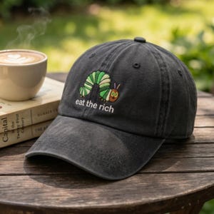 Könnte beinhalten: Dunkelgraue Baseballkappe mit gebogenem Schirm. Die Kappe hat ein gesticktes grünes und gelbes Raupenmuster mit dem Text "eat the rich". Die Kappe liegt auf einem Holztisch.