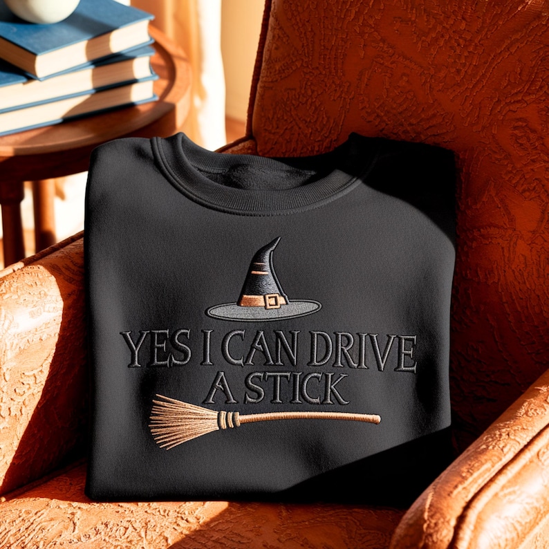 May include: A black crewneck sweatshirt featuring an embroidered design of a witch hat and a broomstick. The text "YES I CAN DRIVE A STICK" is embroidered in black lettering between the hat and the broomstick, centred on the chest.