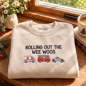 May include: A beige crewneck sweatshirt featuring dark blue embroidered text that reads ROLLING OUT THE WEE WOOS. Below the text are three small embroidered emergency vehicles: a white ambulance, a red fire truck, and a black and white police car.